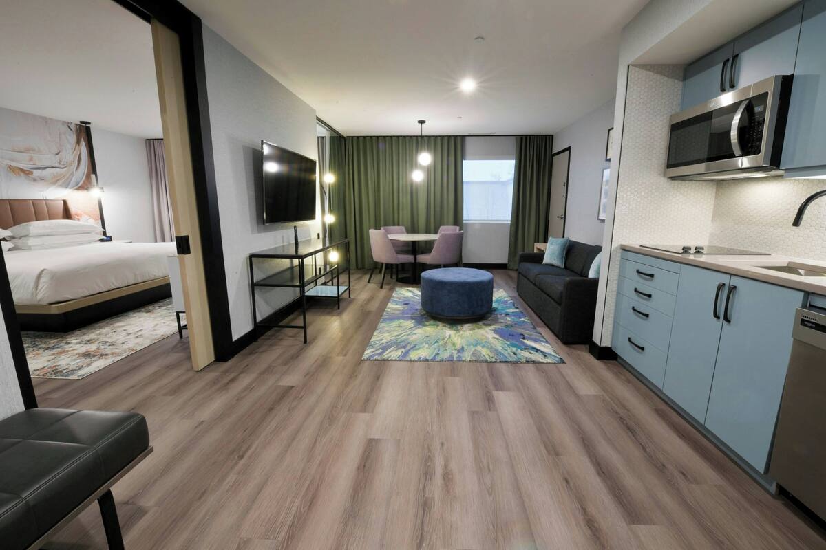A modern hotel room with a bed, a seating area with a table and chairs, a kitchenette, and a large window.