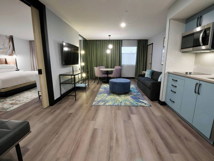 A modern hotel room with a bed, a seating area with a table and chairs, a kitchenette, and a large window.