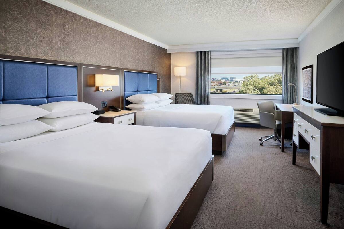 A hotel room with two beds, a desk, a chair, a TV, and a window with a city view.
