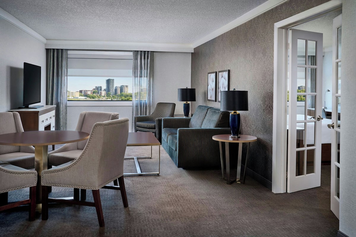 A hotel room with a city view, a sofa, a chair, a small table, a TV, and a door.