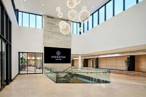 A spacious hotel lobby with a modern design, featuring a large digital sign for Sheraton Laval Hotel, a glass staircase, and a view of the outdoor seating area. - Sheraton Laval Hotel (Laval)