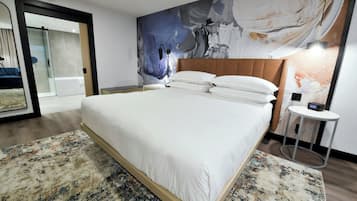 A hotel room with a large bed, a modern headboard, a large abstract painting on the wall, a small round table with a lamp, and a glass door leading to a balcony.