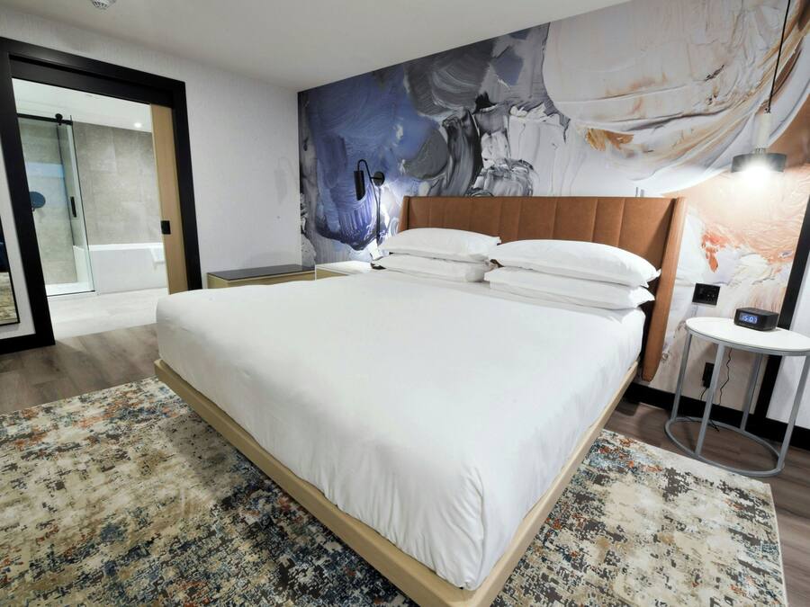 A hotel room with a large bed, a modern headboard, a large abstract painting on the wall, a small round table with a lamp, and a glass door leading to a balcony.