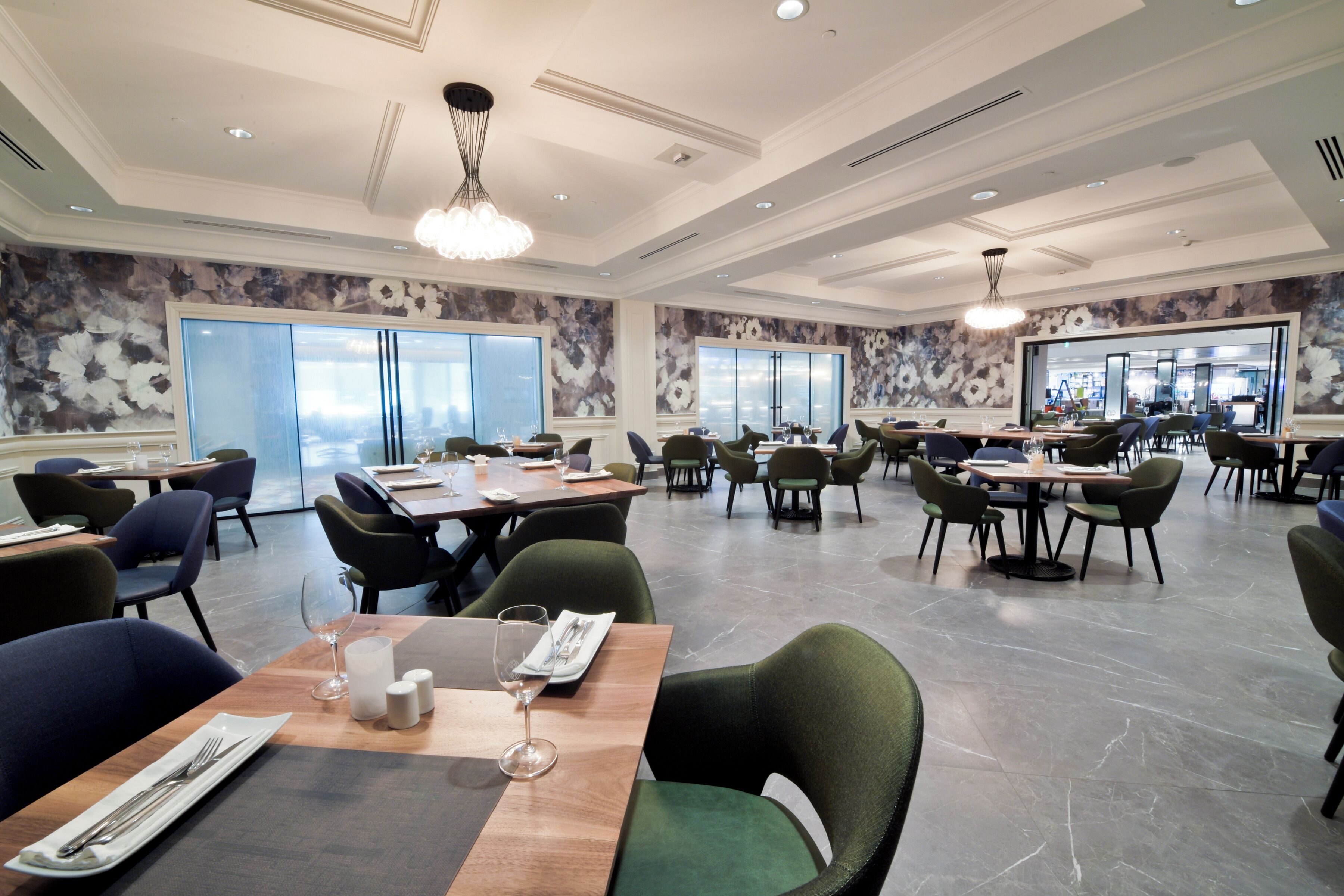 A spacious restaurant with high ceilings, modern chandeliers, and large windows offering views of the city. Tables are neatly arranged with chairs, and the floor is made of polished stone.