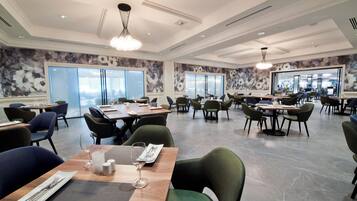 A spacious restaurant with high ceilings, modern chandeliers, and large windows offering views of the city. Tables are neatly arranged with chairs, and the floor is made of polished stone.