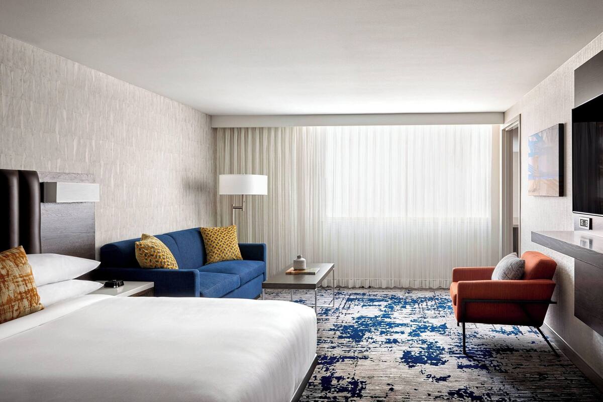 Une chambre d’hôtel avec un grand lit, un canapé bleu, un fauteuil orange, une petite table et un téléviseur fixé au mur.
