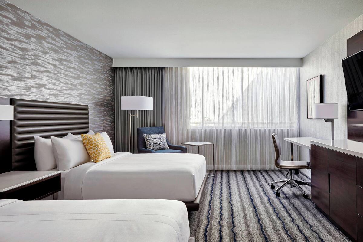 Une chambre d’hôtel avec deux lits, un bureau, une chaise, une télévision et une fenêtre avec des rideaux.