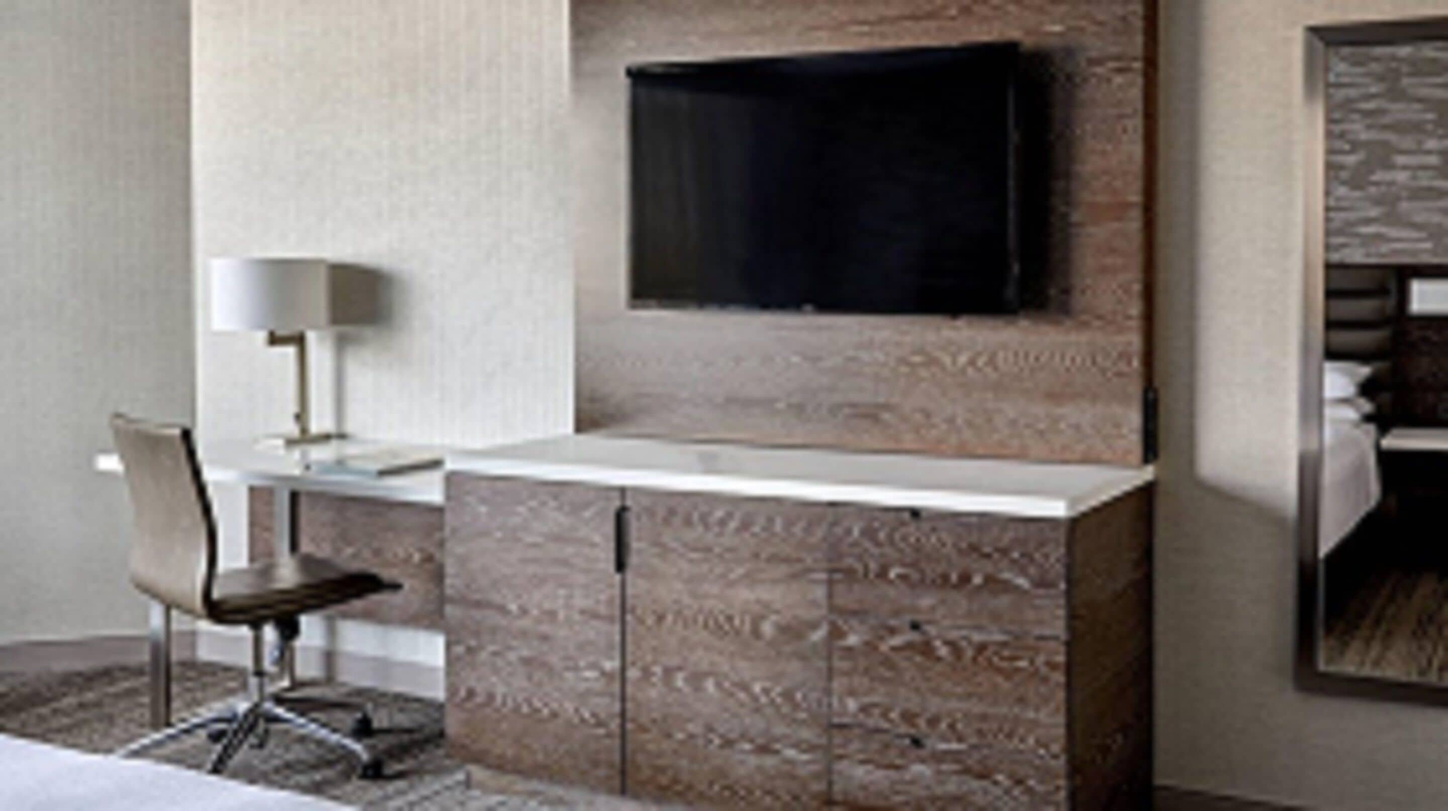 Une chambre d’hôtel moderne dotée d’un téléviseur à écran plat fixé à un mur texturé, d’un bureau avec une chaise et d’un grand miroir.
