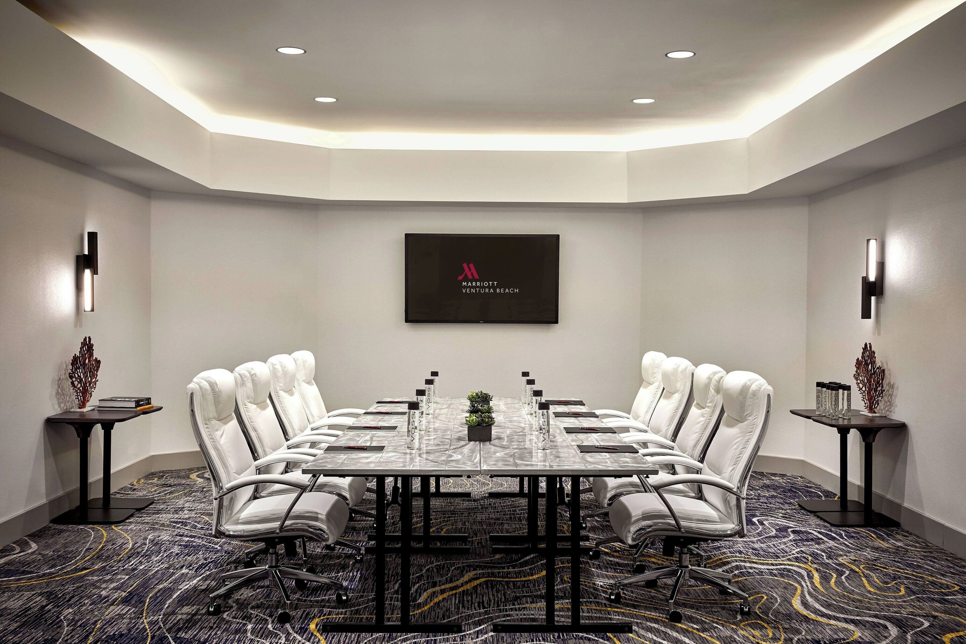 A conference room with a long table, white leather chairs, and a logo on the wall.