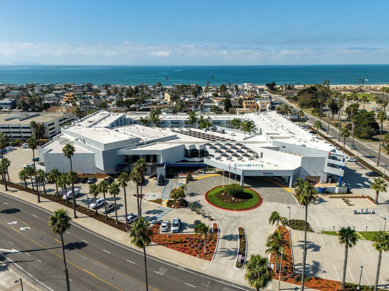 A modern hotel with a central courtyard, surrounded by parking and palm trees.