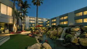 Un hotel moderno con un patio ajardinado que presenta una cascada pequeña, palmeras y una piscina.