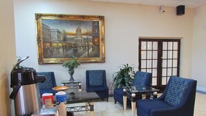 Una habitación con un cuadro grande enmarcado, dos sillones azules, una cafetera, una mesa con un tazón y una taza, y una planta en maceta.