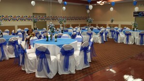 Un salón de banquetes con mesas redondas cubiertas con manteles azules, sillas blancas adornadas con cintas azules y globos.