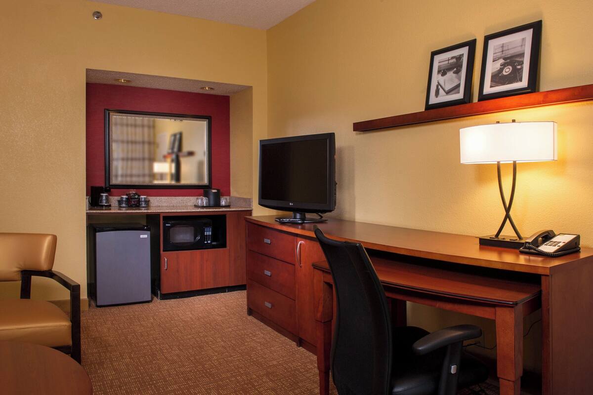 Habitación de hotel con kitchenette, escritorio con computadora y lámpara, una silla y un refrigerador pequeño.