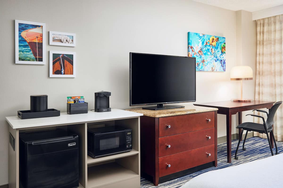 A hotel room with a flat-screen TV, a coffee maker, a microwave, a mini-fridge, a desk with a lamp, and artwork on the wall.