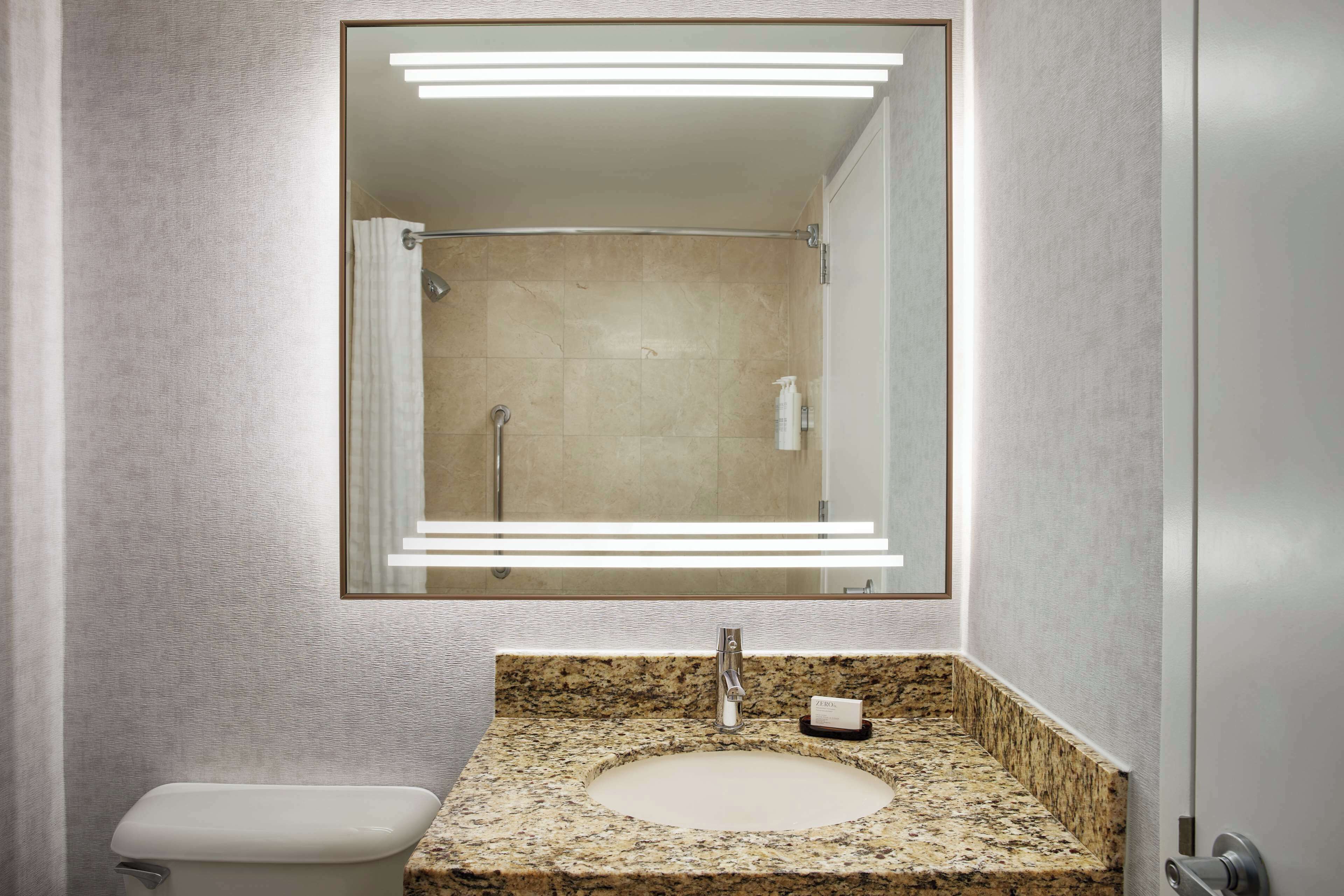 Um banheiro com bancada de granito, pia, espelho com iluminação LED, vaso sanitário e área de chuveiro.