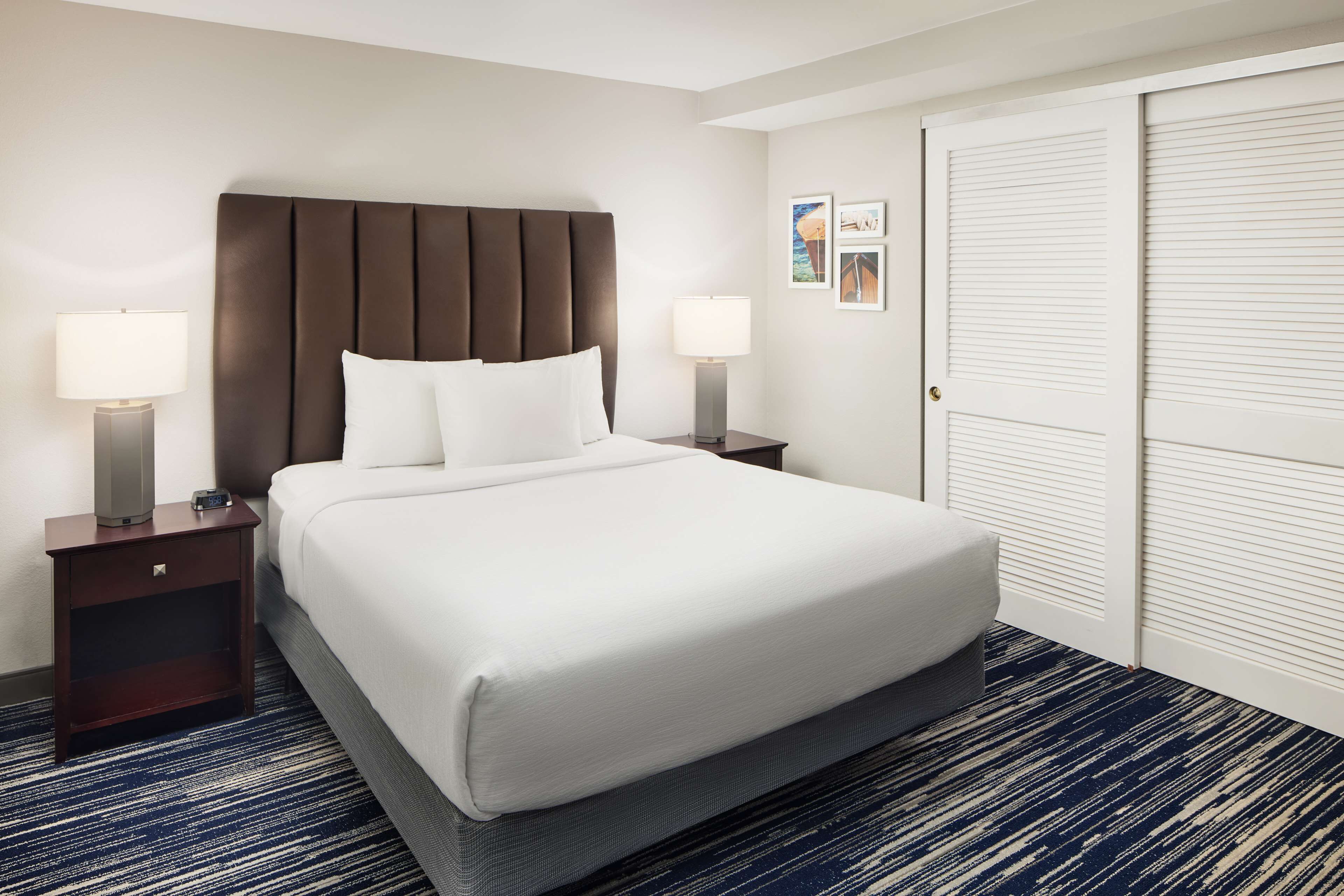 A hotel room with a large bed, two bedside tables with lamps, a dark blue carpet, and a white door with louvered panels.
