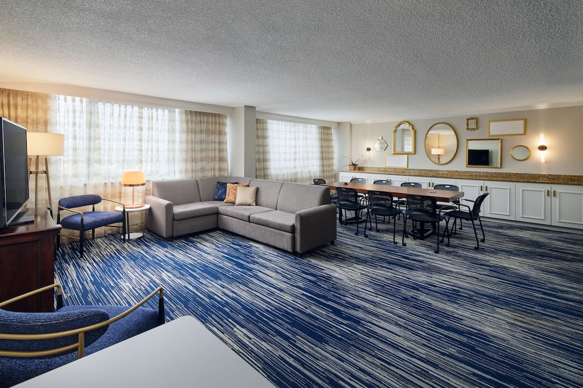 A hotel room with a sectional sofa, a dining table with chairs, a TV, and a large window with curtains.