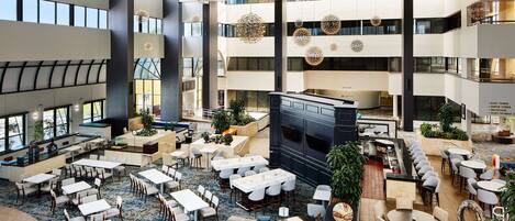 Un amplio vestíbulo de hotel con varias zonas de comedor, un bar central y un gran atrio con luz natural.