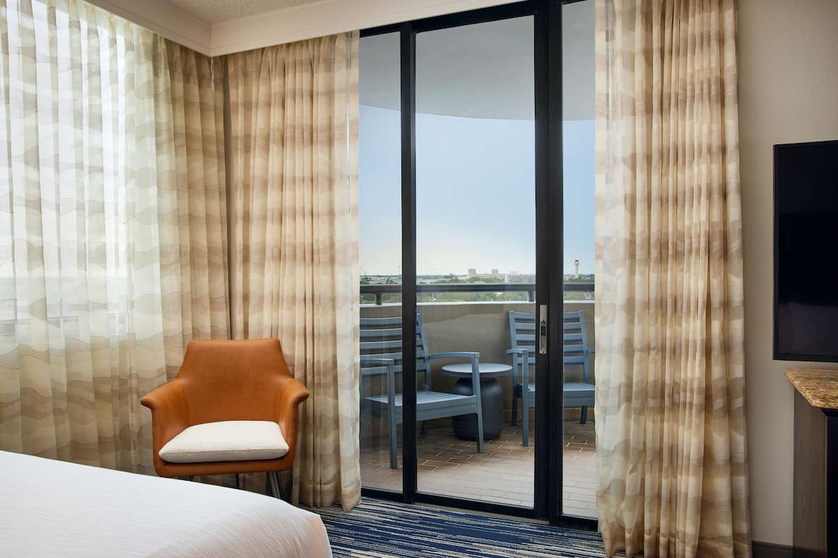 A hotel room with a bed, a chair with a cushion, a glass door leading to an outdoor seating area with chairs and a table, and a flat-screen TV mounted on the wall.