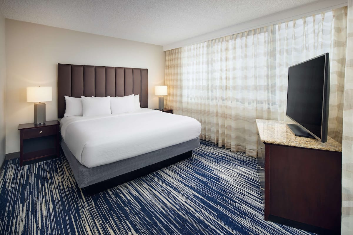 A hotel room with a large bed, two bedside tables with lamps, a flat-screen TV on a wooden console, and a patterned carpet.