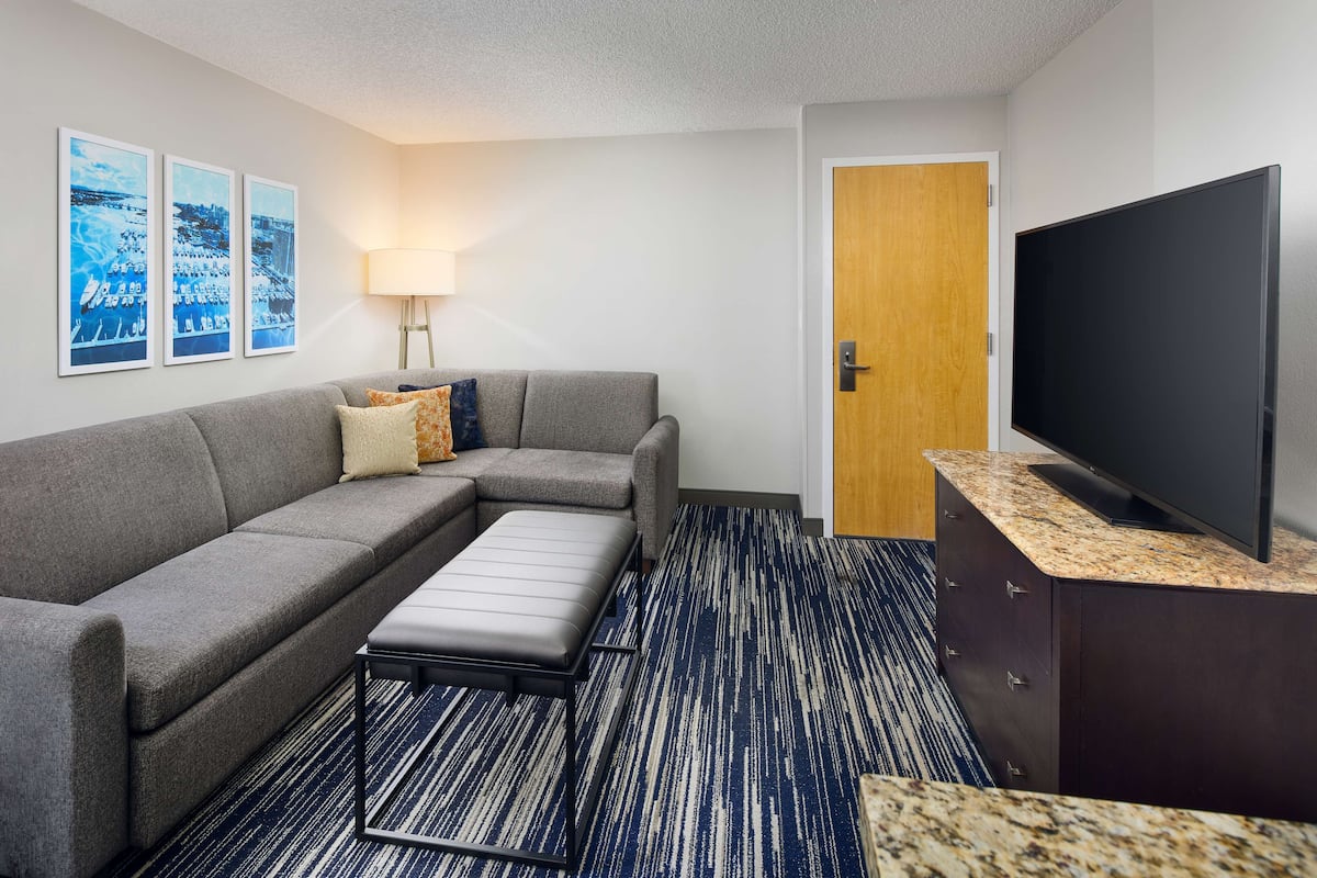 A modern hotel room with a grey sectional sofa, a small ottoman, a flat-screen TV, a wooden door, and a wall-mounted lamp.