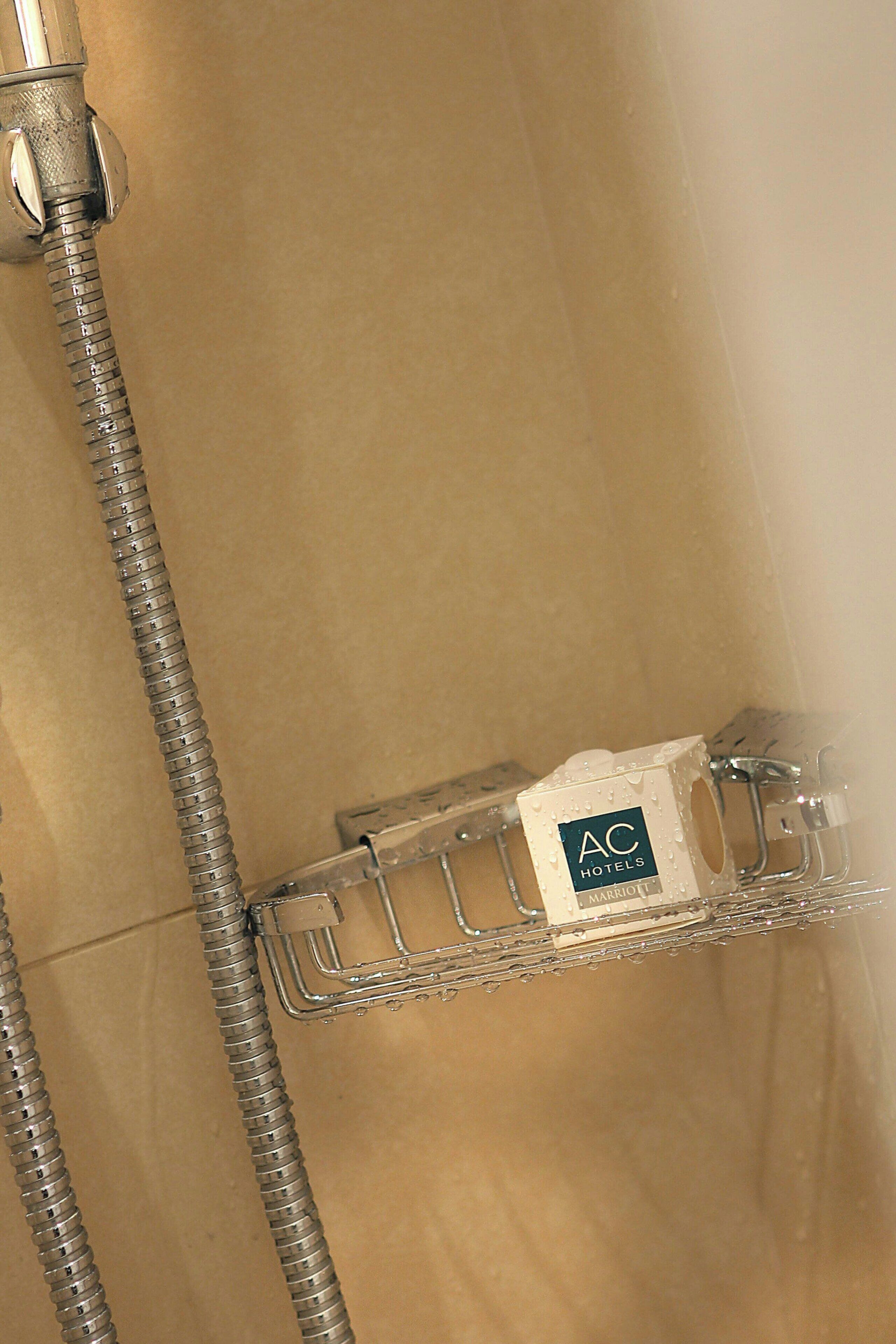 A shower area with a metal hose and a clear plastic shelf with a Marriott AC Hotels logo.