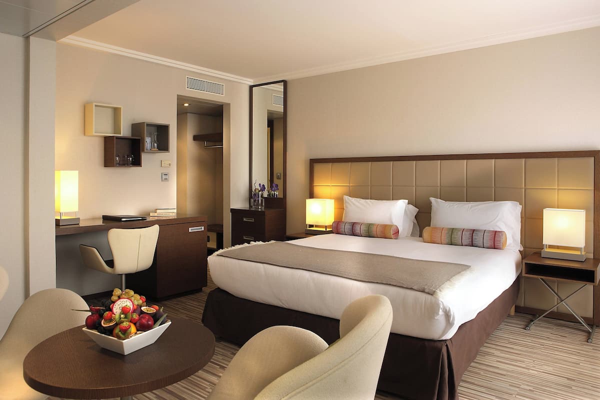 Ein Hotelzimmer mit einem großen Bett, einem Schreibtisch mit Stuhl, einem kleinen Tisch mit Obst und einem Spiegel an der Wand.