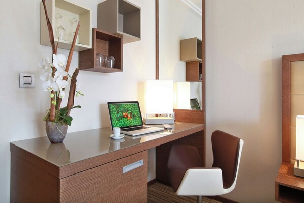 Una habitación de hotel con un escritorio de madera, una laptop, una silla, una orquídea en maceta y un espejo.