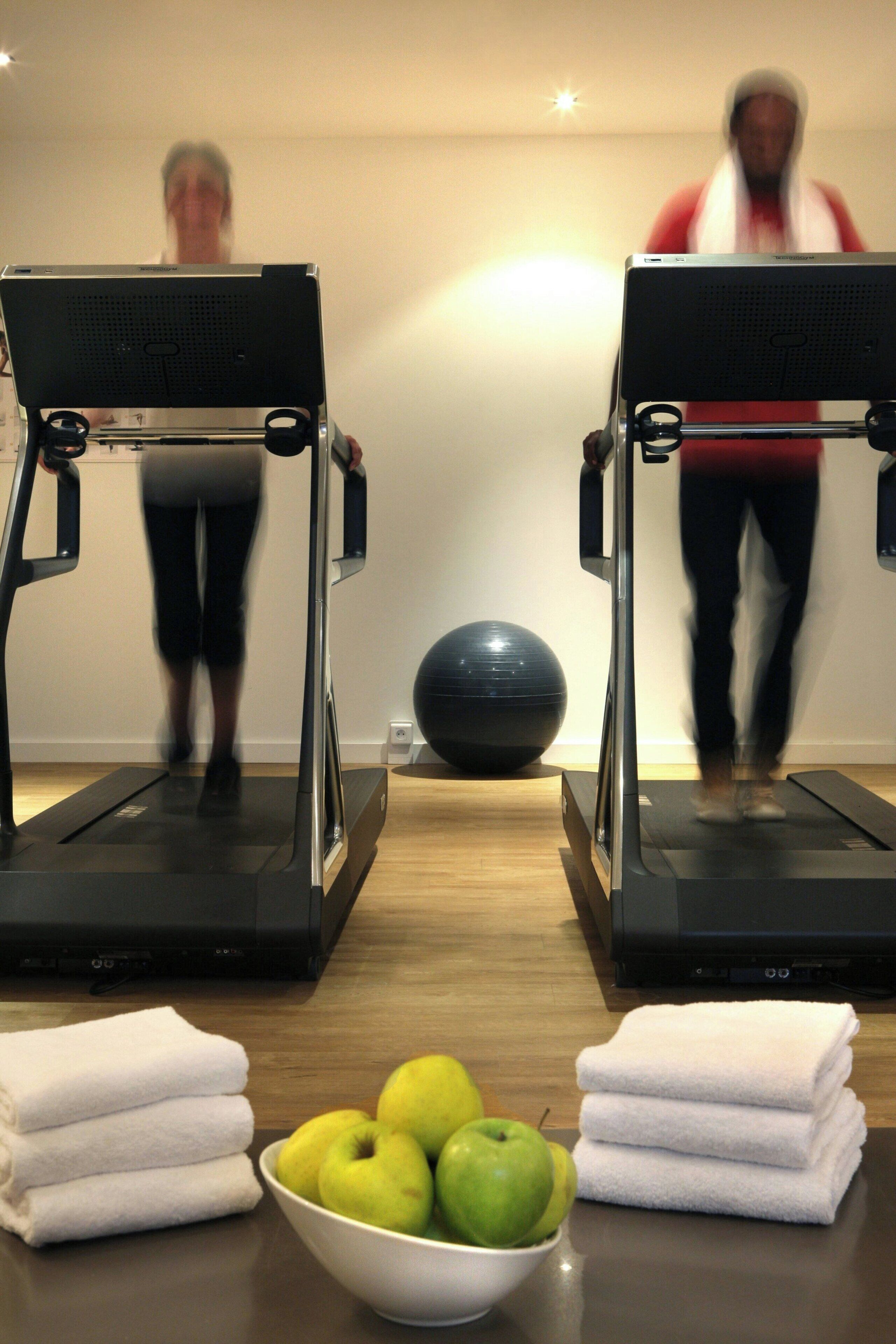 Two exercise machines with a black exercise ball and a bowl of green apples on a wooden floor.