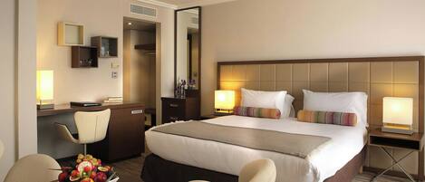 A hotel room with a bed, bedside lamps, a desk with a chair, a small table with fruit, and a mirror.