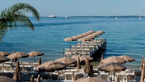 A beachside area with rows of sun loungers and umbrellas, a pier extending into the water, and boats in the background.