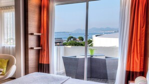 A hotel room with a bed, a chair, a small table, and a balcony with a view of the sea and distant mountains.