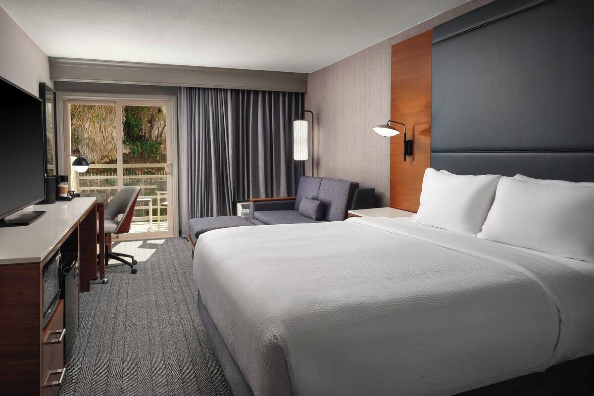 Une chambre d’hôtel avec un grand lit, un bureau, une chaise, une télévision et une fenêtre avec des rideaux.