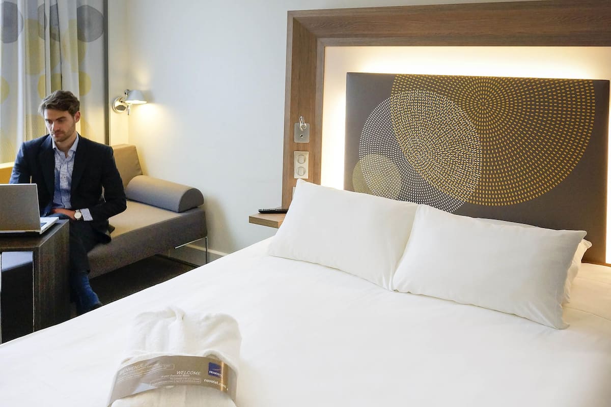 A hotel room with a bed, a desk with a laptop, a chair, a wall art piece, and a welcome towel.