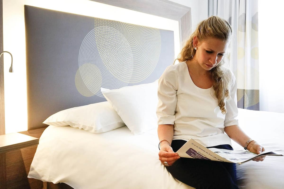 Une personne est assise sur un lit dans une chambre d’hôtel, avec une tête de lit moderne et une grande œuvre d’art abstraite accrochée au mur.