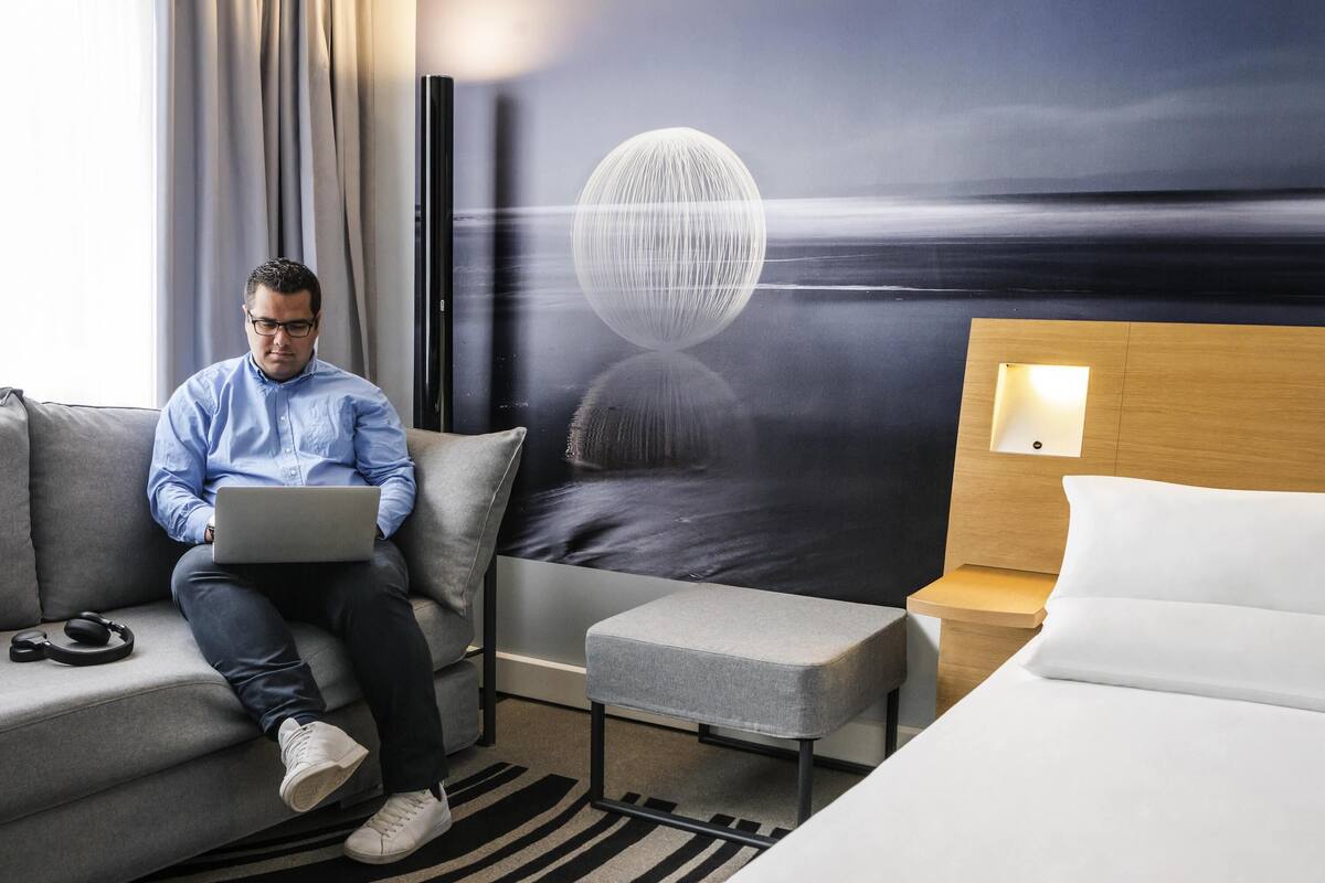 A modern hotel room with a sofa, a bed, a small table, and a wall with a large artwork.
