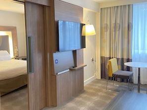 A hotel room with a bed, a TV mounted on a wooden panel, a desk with a lamp, a chair, and a small table.