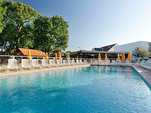 A swimming pool with lounge chairs and umbrellas, surrounded by trees and buildings. - Novotel Paris Saclay (Saclay)