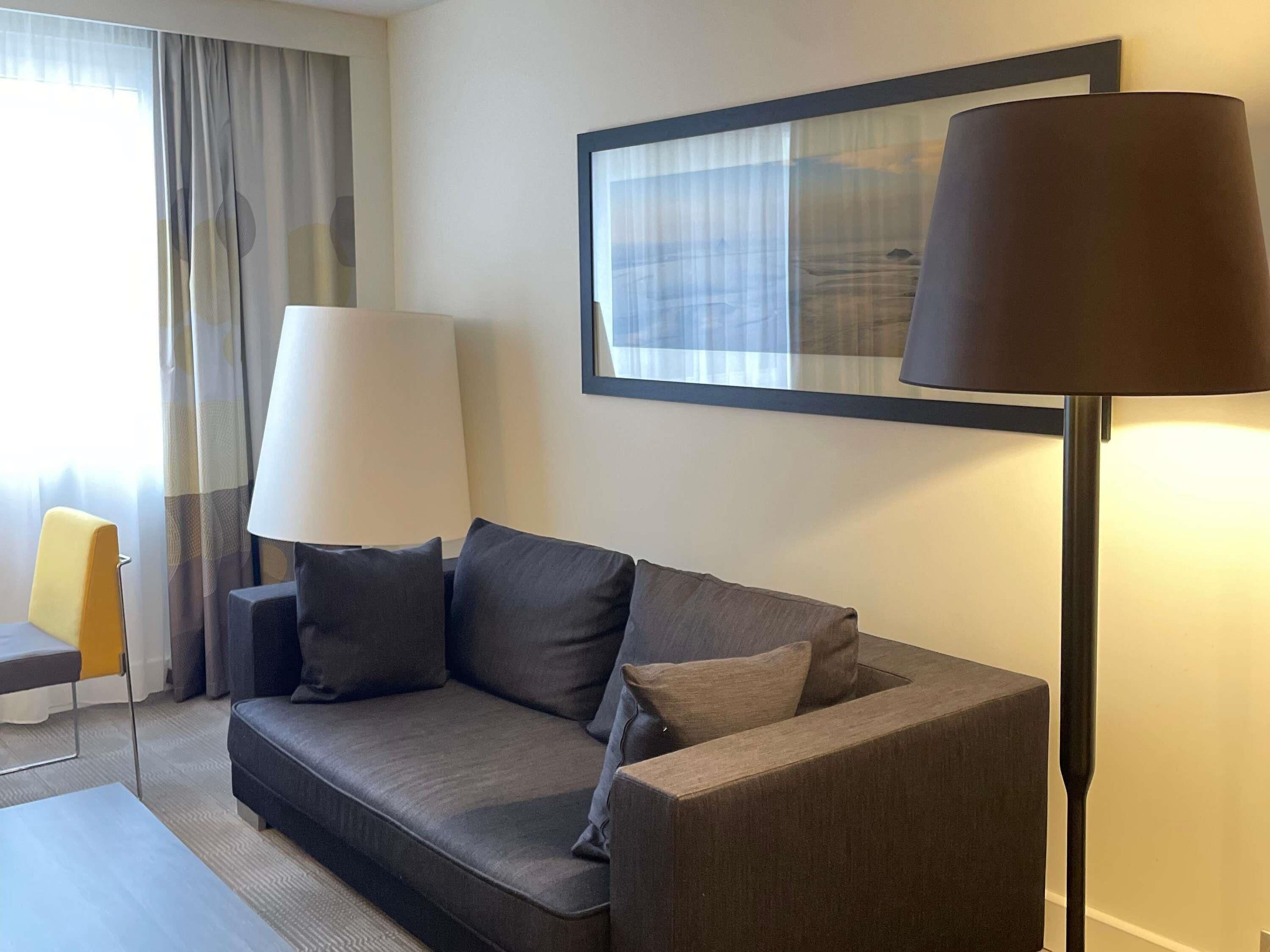 A modern hotel room with a grey sofa, two lamps, a framed artwork on the wall, and a window with curtains.