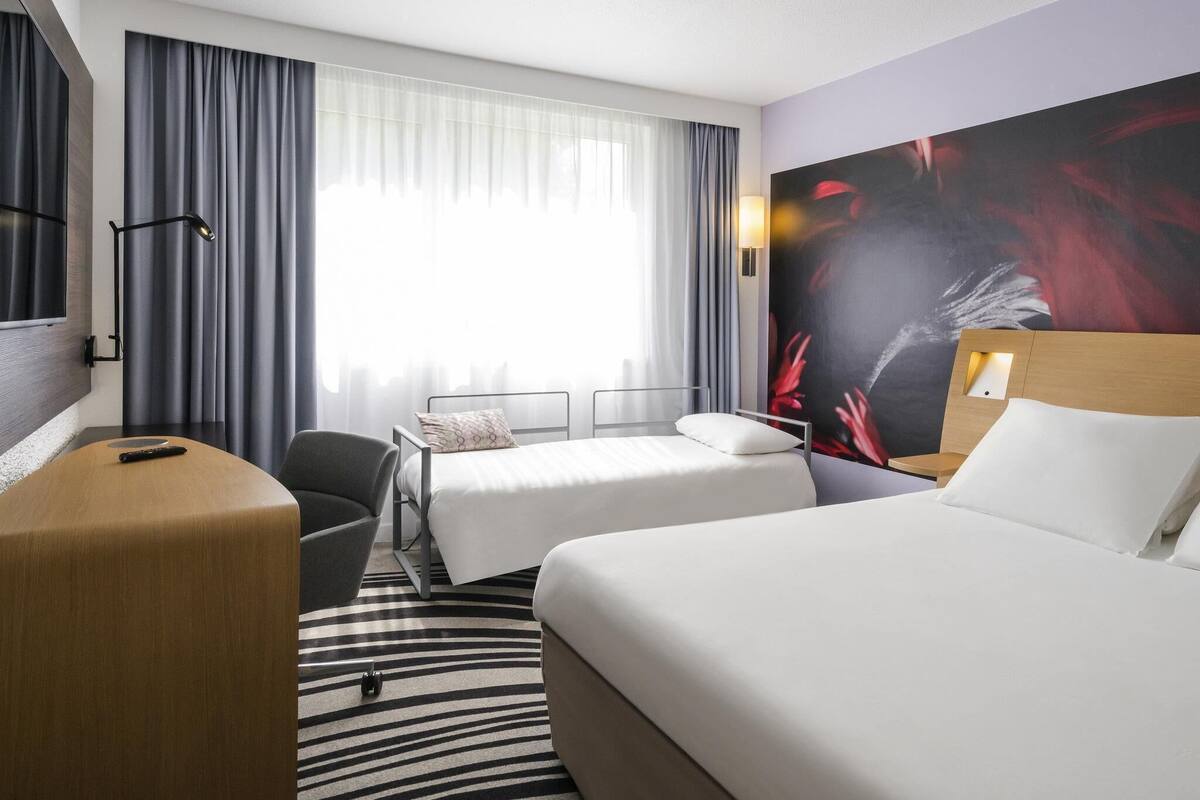 Ett hotellrum med en säng, ett skrivbord, en stol, en TV och ett stort fönster med gardiner.