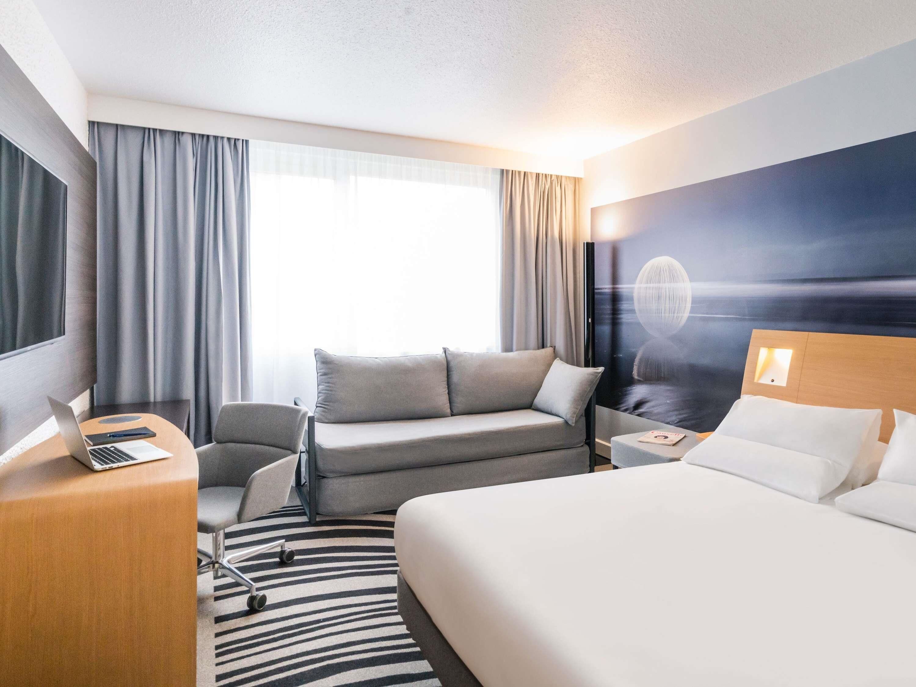 Une chambre d’hôtel avec un lit, un canapé, un bureau avec un ordinateur portable, une chaise et une grande fenêtre avec des rideaux.