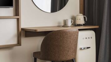 Una moderna camera d'hotel con una scrivania in legno, uno specchio rotondo, una macchina per il caffè, un piccolo frigorifero e una sedia marrone.