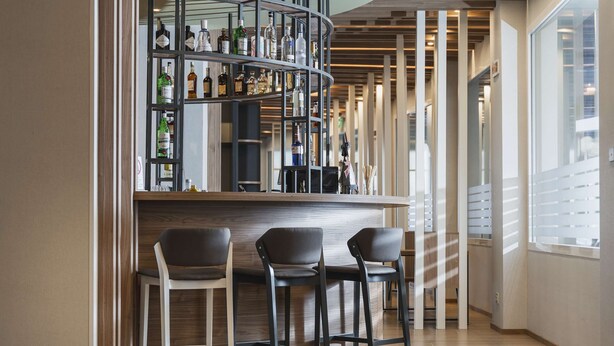 A bar area with wooden ceiling beams, a curved bar counter, and a selection of bottles. There are three chairs in front of the bar. - Hilton Garden Inn Padova City Centre (Padova)