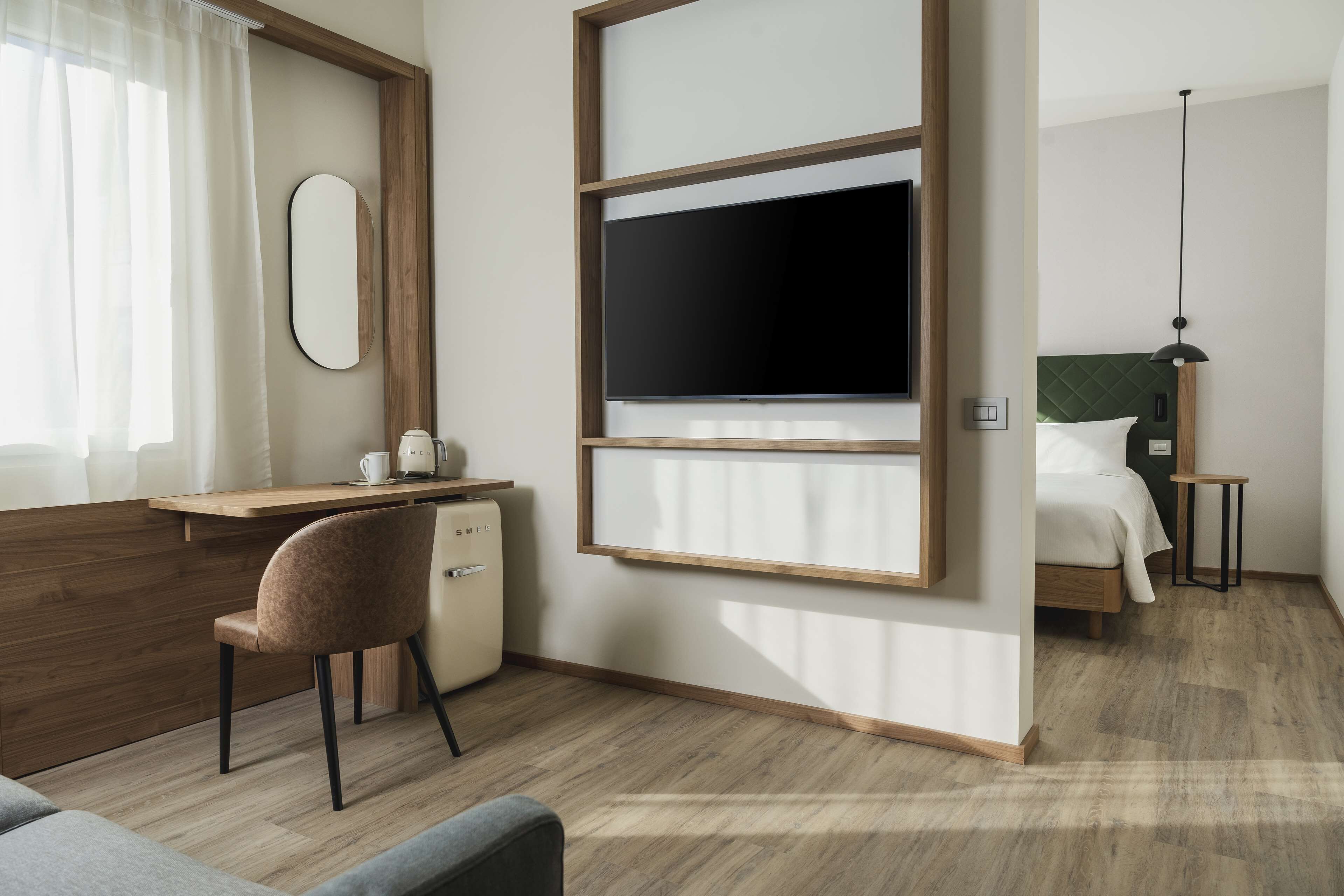 A modern hotel room with a wooden desk, a chair, a bed with a green headboard, and a TV mounted on the wall.