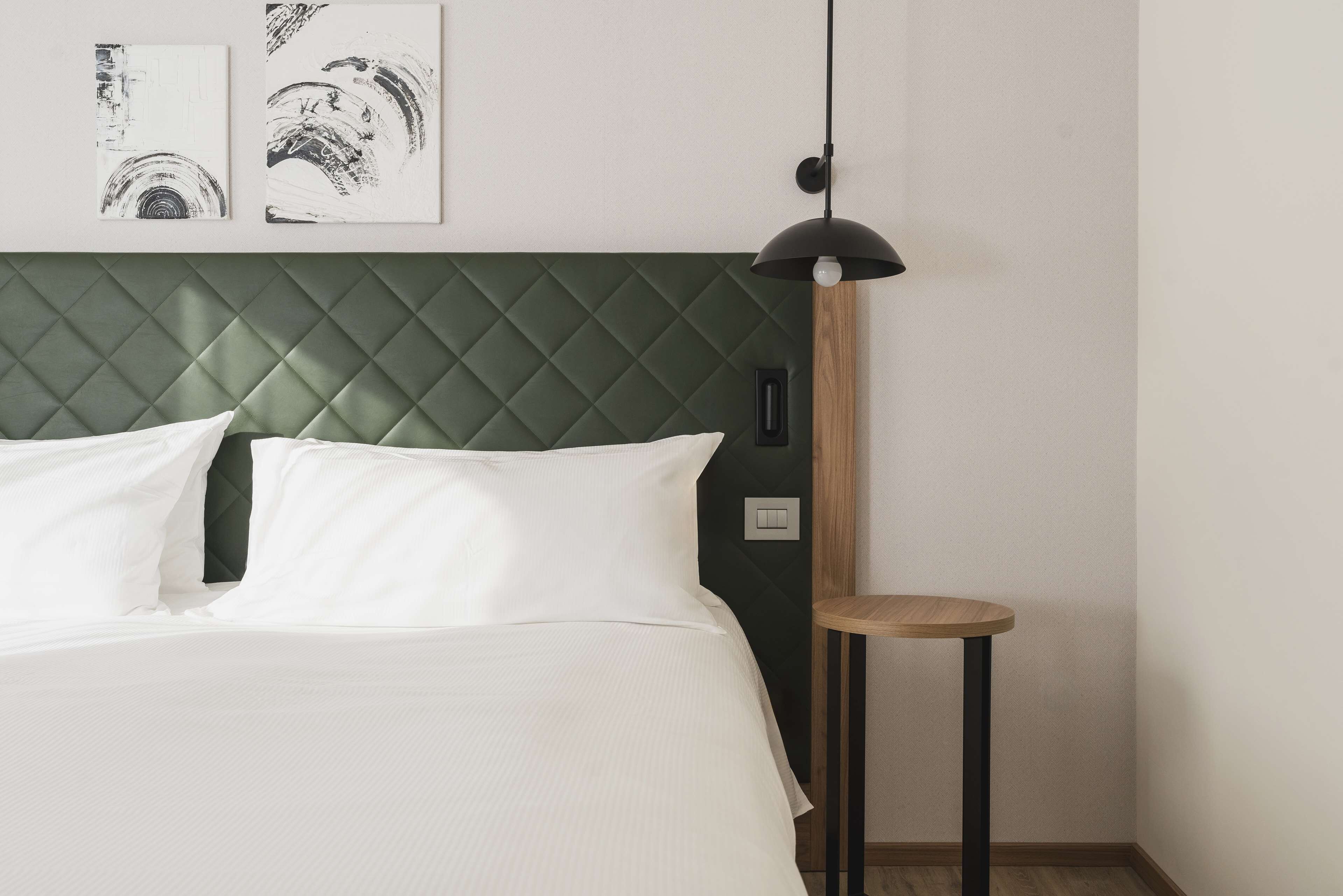 A neatly made bed with white bedding, a green quilted headboard, and two black-and-white artwork pieces on the wall.