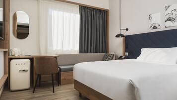 A hotel room with a bed, a desk with a refrigerator, a chair, a window with curtains, a bedside lamp, and artwork on the wall.