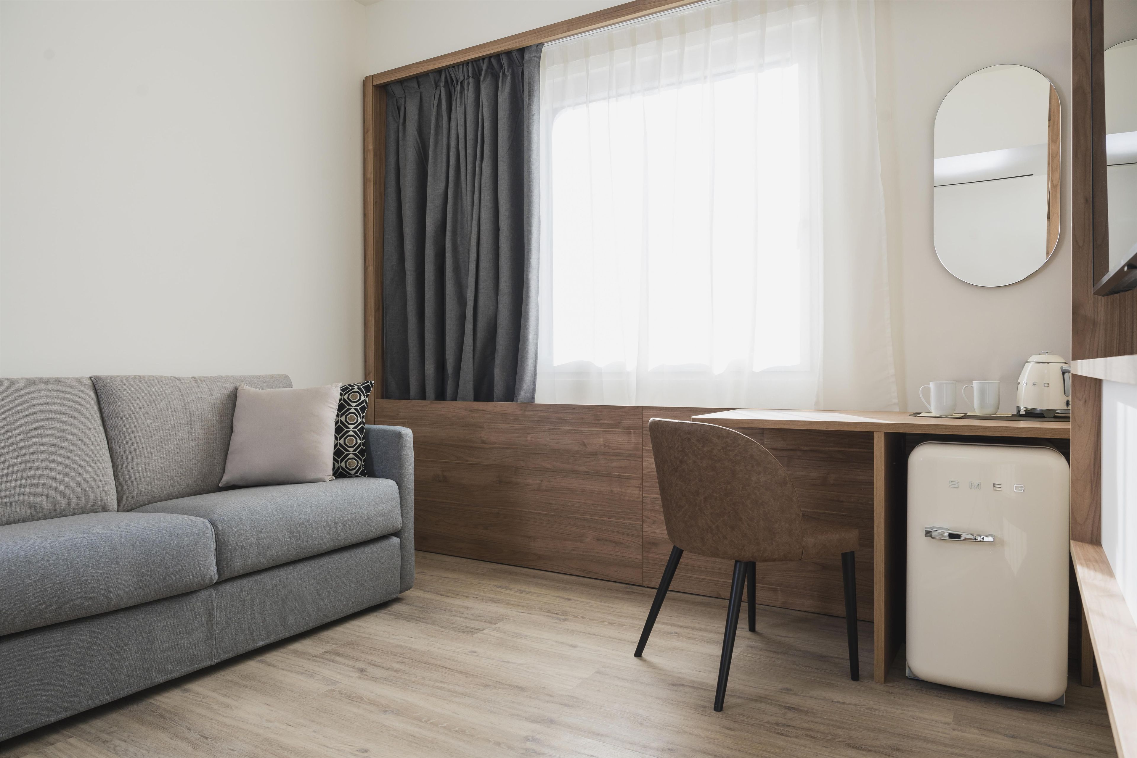 Une chambre d’hôtel moderne avec un canapé gris, un bureau en bois, un petit réfrigérateur et une grande fenêtre avec des rideaux.