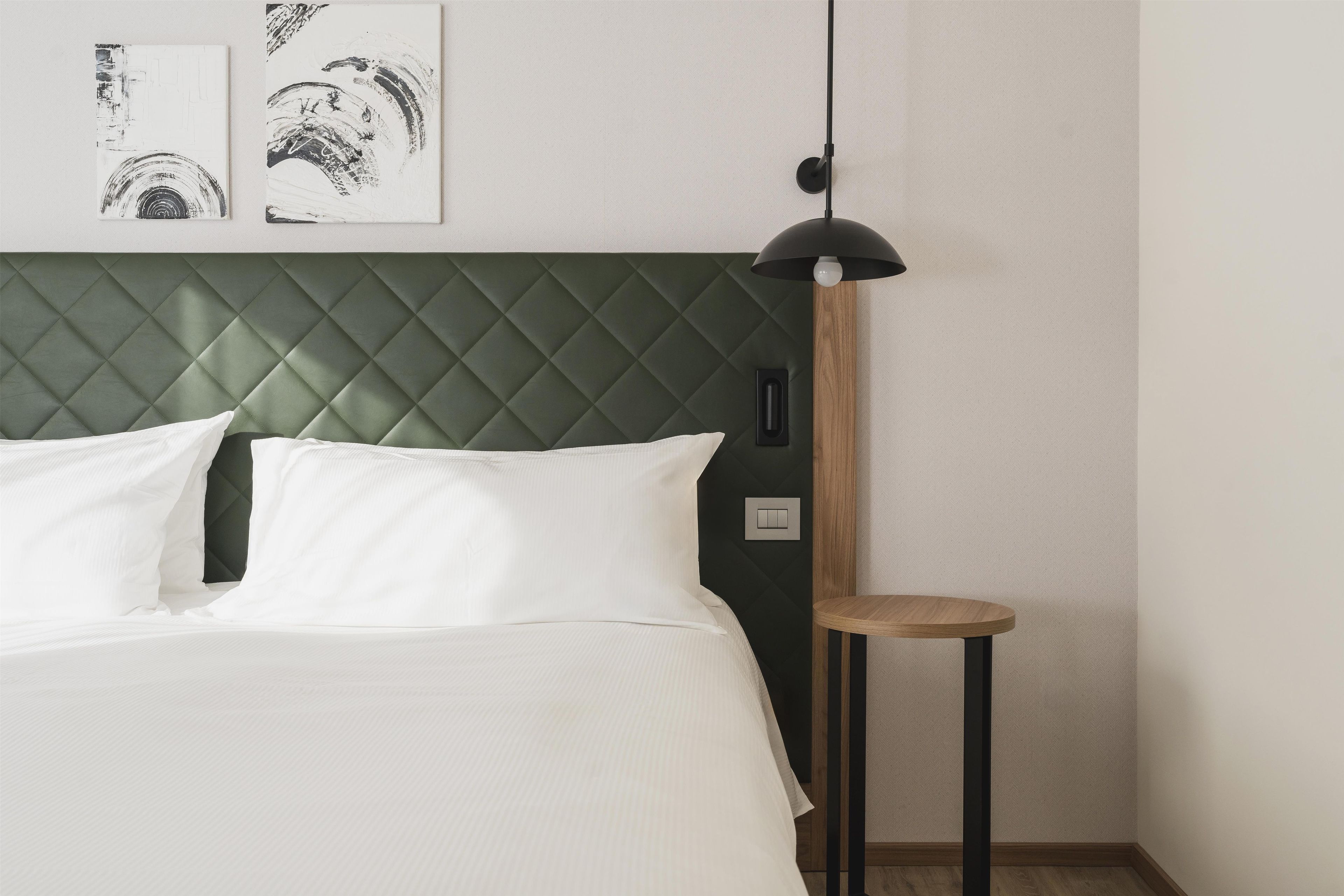 Una cama con ropa blanca, un cabecero acolchado verde y una mesita de noche con una lámpara.