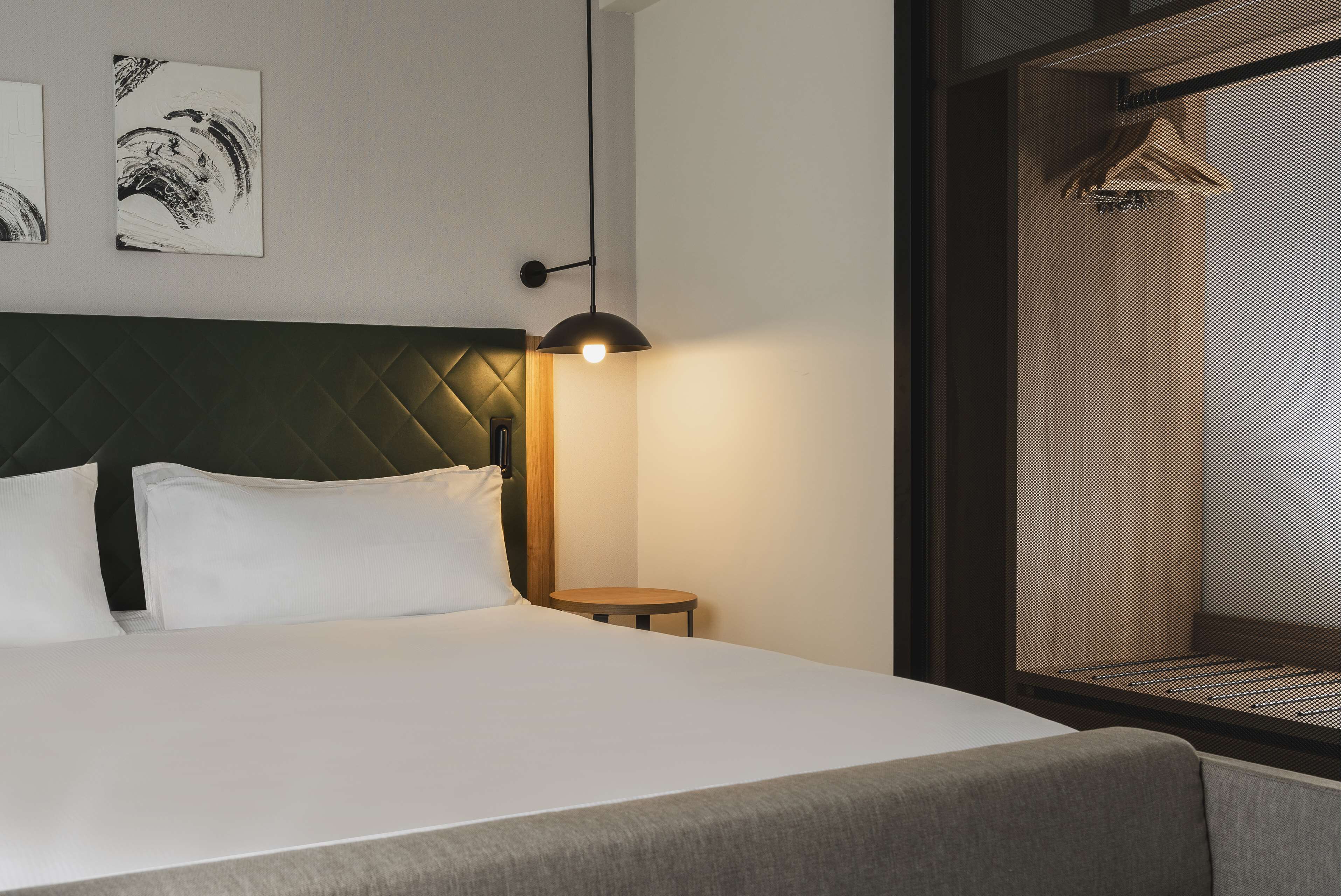 A hotel room with a bed, a round table, a wall-mounted lamp, and artwork.